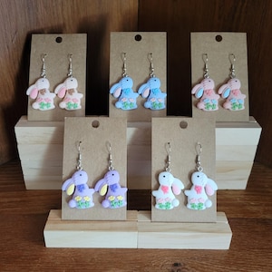 Puede incluir: Seis pares de pendientes colgantes con dijes de conejo con detalles florales. Los conejos son de color rosa, azul, morado y blanco con detalles florales a juego. Los pendientes se muestran en soportes de madera.