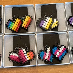 May include: Eight pairs of heart-shaped earrings made with colorful perler beads. Each pair features a different pride flag design, including bisexual, pansexual, transgender, non-binary, lesbian, gay, asexual, and genderqueer.