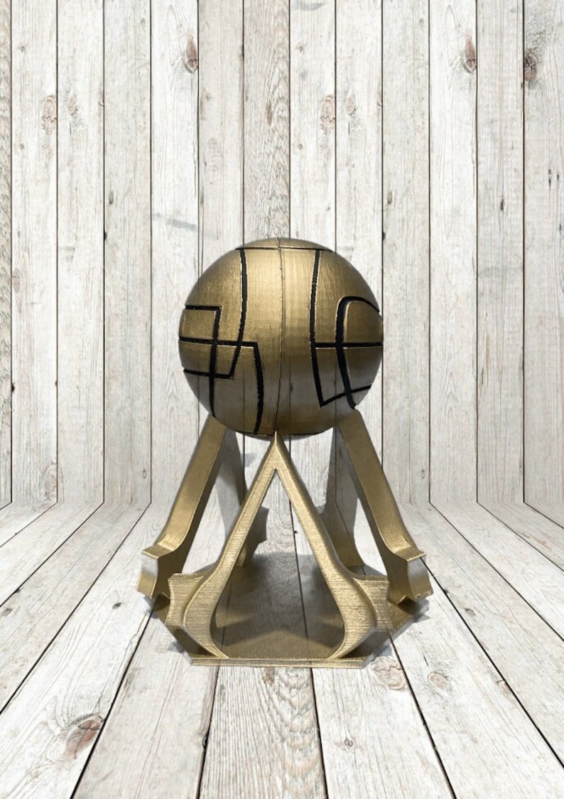 May include: A gold-colored decorative sphere with black geometric designs, mounted on a matching gold stand. The stand features a stylized, three-legged design. The object is set against a light wood-grain background.