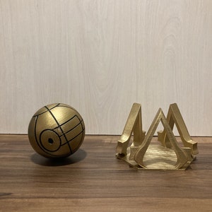 May include: A gold-colored, spherical object with black lines and a geometric design, next to a gold-colored, stylized stand. The stand has a hexagonal base and a repeating triangular design. Both items appear to be 3D-printed.