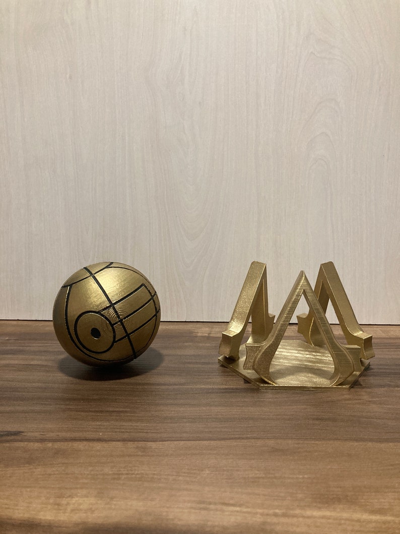 May include: A gold-colored, spherical object with black lines and a geometric design, next to a gold-colored, stylized stand. The stand has a hexagonal base and a repeating triangular design. Both items appear to be 3D-printed.