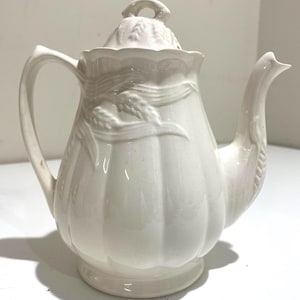 May include: A white ceramic teapot with a lid, handle, and spout. The teapot has a ribbed body and is decorated with embossed floral and ribbon designs. The handle and spout have decorative details. The teapot is on a white surface.