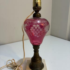 Puede incluir: Una lámpara de mesa vintage con una pantalla de cristal rojo rubí grabada con un diseño de uvas. La lámpara tiene una base de latón y una base cuadrada de mármol. La lámpara está apagada y tiene un cable eléctrico beige.