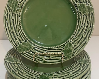 Puede incluir: Un juego de seis platos de cerámica verdes. Cada plato presenta un diseño en relieve de ramas, cuerda y hojas de roble alrededor del borde. Los platos están apilados en un soporte de metal dorado.