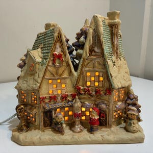 May include: A ceramic Christmas village scene with two buildings, snow-covered roofs, and lit windows. The buildings are decorated with red bows and garlands. Small figurines of people and a snowman are in front of the buildings.