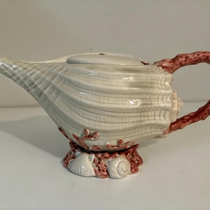 May include: A ceramic teapot shaped like a seashell, with a white and gray body and a coral-colored handle and base. The teapot has a lid and is decorated with coral and shell details.