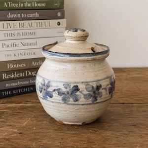 May include: A round, beige ceramic jar with a matching lid, featuring a hand-painted horizontal band of blue flowers and thin blue lines. The jar has a speckled texture, a slightly flared rim, and a small notch in the lid for a spoon or utensil.
