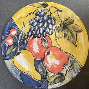 May include: A hand-painted ceramic plate with a fruit design. The plate has a yellow background with a blue border and features grapes, apples, and pears in red, yellow, and blue. A decorative item for the kitchen.