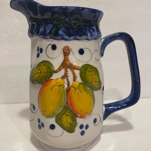 May include: A white ceramic jug with a blue handle and rim. The jug features a painted design of lemons with green leaves and a brown stem.