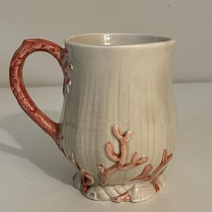Puede incluir: Taza de cerámica color crema con diseño de arrecife de coral. El asa y los detalles de coral son de color naranja rojizo. La taza tiene una superficie texturizada y una base ancha.