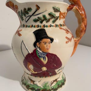 May include: A white ceramic jug with a fox-shaped handle. The jug is decorated with a hunting scene featuring a man in a red jacket and a black top hat, holding a whip. There are also images of dogs and trees.