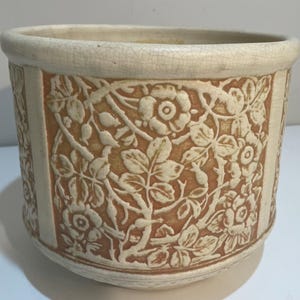 Puede incluir: Jardinera de cerámica color crema con diseño floral en relieve. La jardinera tiene una forma ovalada ancha con un borde texturizado blanquecino. El cuerpo de la jardinera tiene un fondo marrón anaranjado cálido con patrones florales y de hojas blancos en relieve.