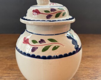 Może przedstawiać: Biały ceramiczny słoik z pokrywką, ozdobiony kwiatowym wzorem w odcieniach czerwieni, fioletu, zieleni i błękitu. Słoik ma falisty brzeg i gałkę na pokrywce.