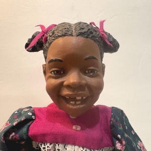 Puede incluir: Una muñeca negra sonriente con el pelo castaño oscuro en coletas atadas con cintas rosas. La muñeca lleva un vestido rosa y verde con estampado floral.