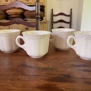 May include: Four off-white ceramic teacups with handles are arranged on a wooden surface. Each cup has a ribbed design and a decorative rope-like trim near the rim. The cups are empty.