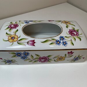 Puede incluir: Caja de pañuelos rectangular blanca con diseño floral. La caja presenta un borde dorado y una abertura ovalada. Las flores son en tonos rosa, amarillo y azul, con hojas verdes. Un artículo decorativo para el hogar.