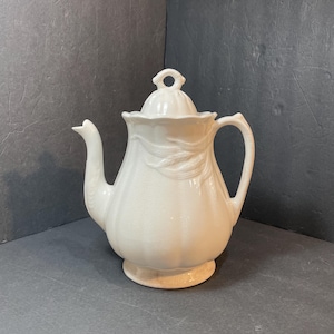 May include: A white ceramic teapot with a lid, spout, and handle. The teapot has a rounded body with a decorative embossed design and sits on a small, circular base. The lid has a loop handle.
