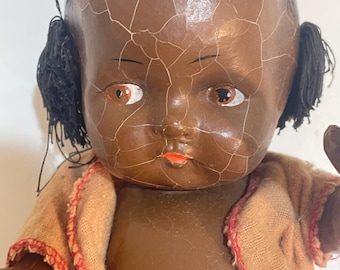 May include: Vintage black doll with brown eyes, black hair, and a cracked, aged appearance. The doll wears a tan jacket with red trim. The doll's face has a painted orange mouth and delicate eyebrows. A collectible item.