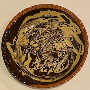 May include: A round ceramic plate with a brown rim and a swirling pattern of black and yellow glaze.