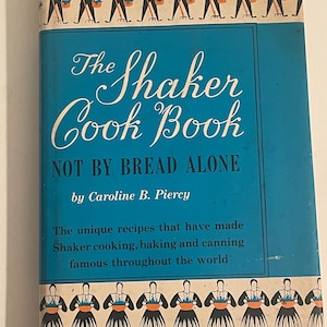 Op de afbeelding: Vintage kookboek getiteld "The Shaker Cook Book" door Caroline B. Piercy. Het boek heeft een turquoise omslag met witte tekst en illustraties van Shaker-figuren. De titel "NOT BY BREAD ALONE" is ook zichtbaar.