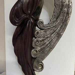 Op de afbeelding: Een bronzen beeld van een vrouw met een vloeiende jurk en een grote, sierlijke vleugel. Het beeld is afgewerkt in donkerbruin en zilver.