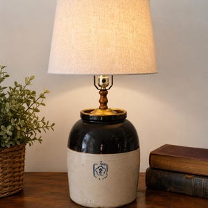 Könnte beinhalten: Eine Tischlampe mit einem zylindrischen Keramikfuß in zweifarbigem Design, dunkelbraun in der oberen Hälfte und cremeweiß in der unteren Hälfte, markiert mit einer blauen Ziffer 2 in einem Blumenemblem. Sie hat einen beigen Stoffschirm in Trommelform.