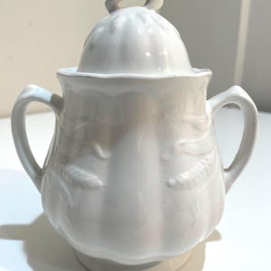 May include: White ceramic sugar bowl with a lid and two handles. The bowl has a rounded shape with embossed floral designs and a small loop handle on the lid. The sugar bowl is a classic design.