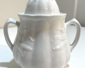 Może przedstawiać: Biała ceramiczna cukiernica z pokrywką i dwoma uchwytami. Miska ma zaokrąglony kształt z wytłoczonymi wzorami kwiatowymi i małym uchwytem pętelkowym na pokrywce. Klasyczny design.