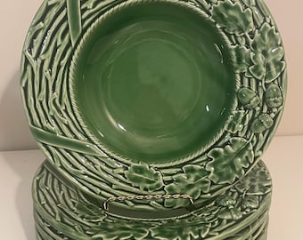 Puede incluir: Una pila de seis platos de cerámica verde con un diseño decorativo. Los platos presentan un borde con un patrón de ramas tejidas y hojas de roble con bellotas. Los platos están apilados en un soporte de metal dorado.