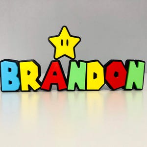May include: A vibrant, 3D-printed name sign spelling "BRANDON" in block letters. The letters are in blue, yellow, red, green, and red. A yellow star with a black outline is positioned above the name, adding a playful touch.