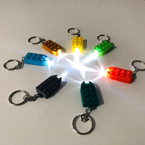 May include: A selection of small, colourful brick-shaped LED keyrings. The keyrings are in various colours, including blue, yellow, green, red, black, and teal. Each keyring is emitting a bright white light, creating a starburst effect.