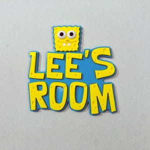 May include: A decorative wall sign featuring a yellow, porous character face above the blue text LEE'S ROOM. The letters are yellow with a porous texture, set against a blue background. The design includes two white eyes and two teeth.