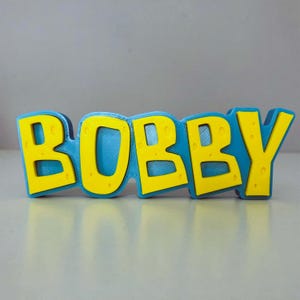 May include: A 3D-printed sign that spells out the name "BOBBY" in bold, yellow letters with a textured surface. The letters are outlined in a vibrant blue, creating a striking contrast. The sign is designed to stand on a flat surface.