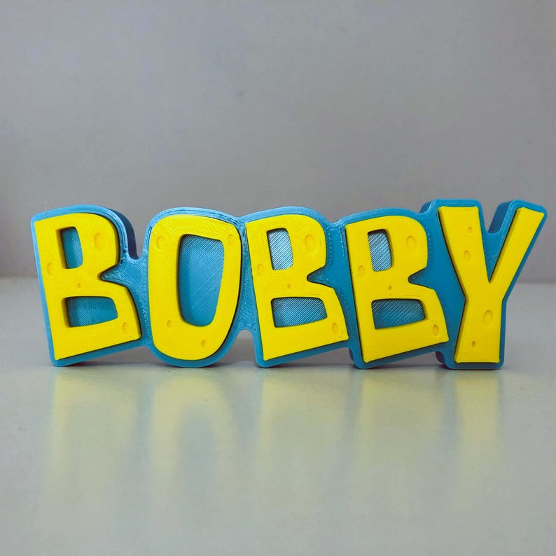 May include: A 3D-printed sign that spells out the name "BOBBY" in bold, yellow letters with a textured surface. The letters are outlined in a vibrant blue, creating a striking contrast. The sign is designed to stand on a flat surface.