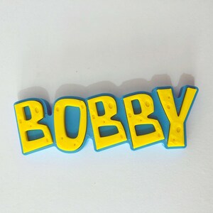 May include: A colorful, three-dimensional nameplate that spells out "BOBBY" in bold, yellow letters with a textured pattern. The letters are outlined in a vibrant blue, creating a playful and eye-catching design. The nameplate is likely a decorative item.