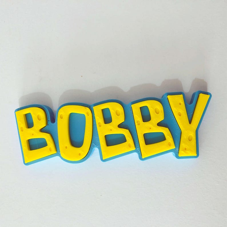 May include: A colorful, three-dimensional nameplate that spells out "BOBBY" in bold, yellow letters with a textured pattern. The letters are outlined in a vibrant blue, creating a playful and eye-catching design. The nameplate is likely a decorative item.