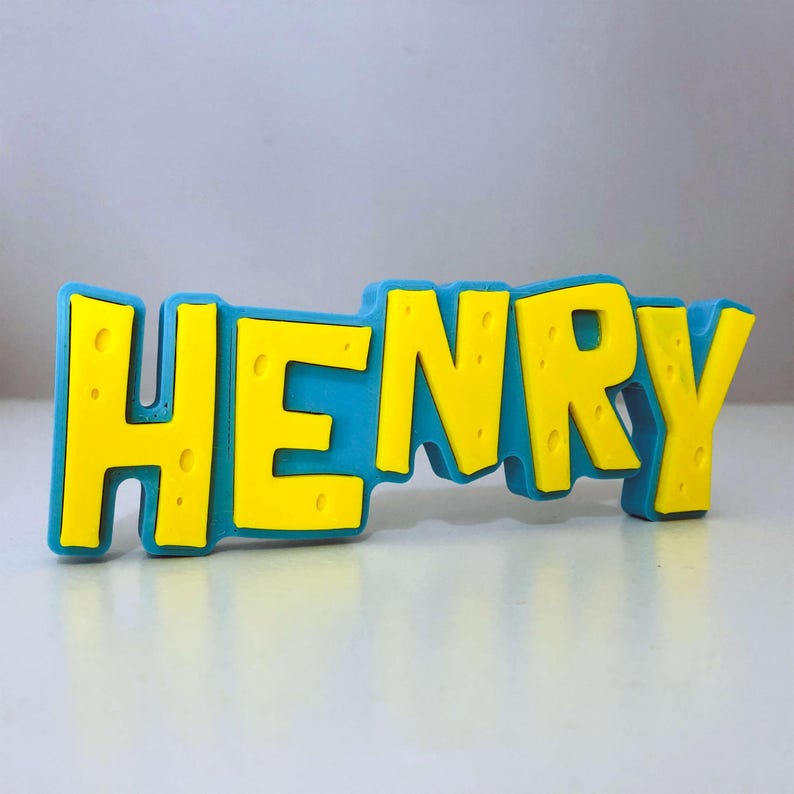 May include: A blue and yellow nameplate that spells out the name "HENRY". The letters are yellow with a textured appearance, set against a blue background. The nameplate is designed in a playful, blocky font.