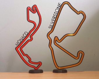 May include: Two black metal race track outlines, one red and one orange, stand on a wooden surface. The red track is labeled "MONACO" and the orange track is labeled "SILVERSTONE".
