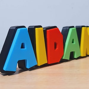 May include: A colourful 3D name sign spelling "AIDAN" with each letter in a different colour: blue, yellow, red, green, and yellow.