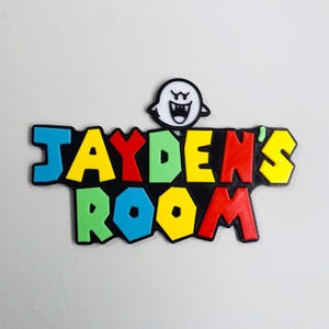 May include: A vibrant sign reading "JAYDEN'S ROOM" in blue, yellow, red, and green, with a black border. A white ghost character sits above the text. Ideal for a child's bedroom, the sign is eye-catching and playful.