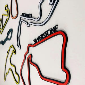 May include: A selection of colourful, three-dimensional race track outlines, including Silverstone and Monaco, mounted on a white surface. The tracks are in black, red, yellow, and orange, with track names in bold, black lettering.