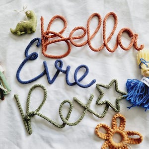 May include: Handmade nursery decor with the names "Bella", "Evie", and "Rex" in orange, blue, and green yarn. Includes a green dinosaur, star, and flower accents. A blue tassel with "Proud of Yourself" text is also present.