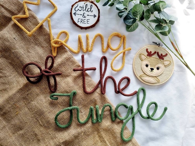 May include: A nursery decor set with the words "King of the Jungle" spelled out in yarn letters. The letters are in different colors: yellow, brown, and green. There is also a small felt deer head with antlers in a wooden hoop.