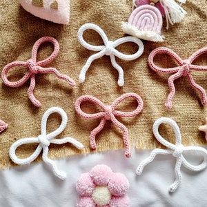 May include: Handmade crochet bows in white and pink, displayed on a textured, tan fabric. The image includes decorative elements such as stars and a flower. These bows are suitable for crafting and embellishments, perfect for adding a touch of charm.