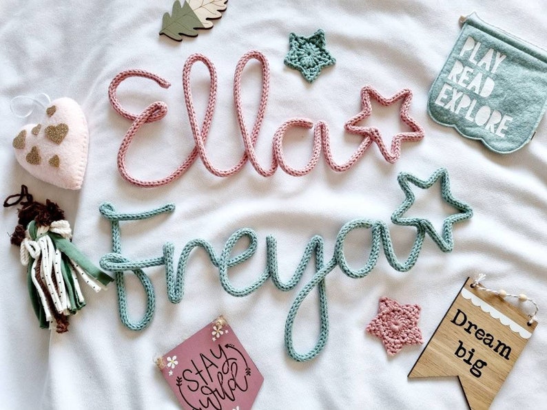 May include: Two knitted names, "Ella" in pink and "Freya" in blue, with star shapes. Other decorations include a heart, a banner with "Play Read Explore", and a wooden sign saying "Dream big". The items are arranged on a white surface.