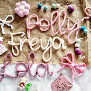 May include: A selection of handmade nursery decorations, including crocheted name signs spelling "Polly", "Freya", and "Lola". The image also features crocheted bows, a flower, a rainbow, and a garland of pom-poms. A small sign reads "Stay Wild".