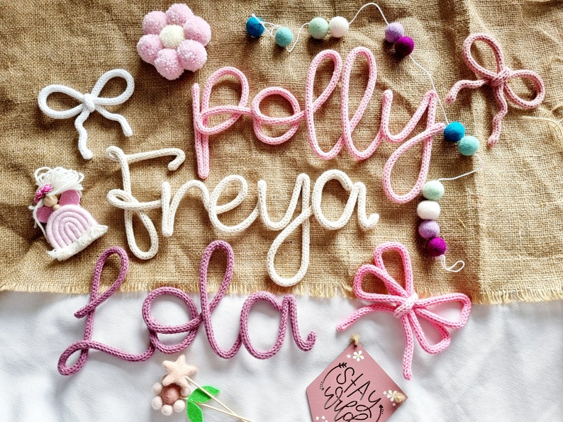 May include: A selection of handmade nursery decorations, including crocheted name signs spelling "Polly", "Freya", and "Lola". The image also features crocheted bows, a flower, a rainbow, and a garland of pom-poms. A small sign reads "Stay Wild".