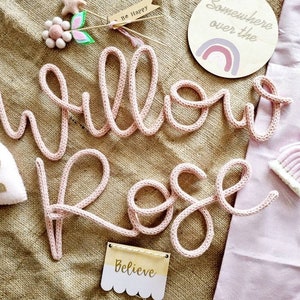 May include: A pink knitted 'Willow Rose' name sign, with decorative elements including a heart, a rainbow, and a wooden sign that says 'Believe'. The sign is displayed on a neutral burlap surface.