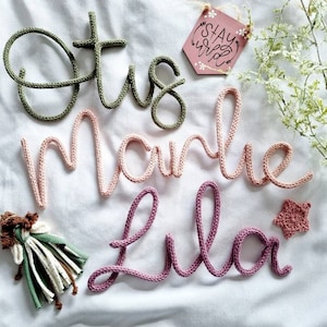 May include: Crochet lettering spelling "Etsy", "Marie", and "Lila" in green, pink, and purple yarn. A small wooden tag with the words "Stay Wild" and a star-shaped crochet embellishment are also present.