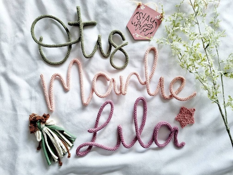 May include: Crochet lettering spelling "Etsy", "Marie", and "Lila" in green, pink, and purple yarn. A small wooden tag with the words "Stay Wild" and a star-shaped crochet embellishment are also present.